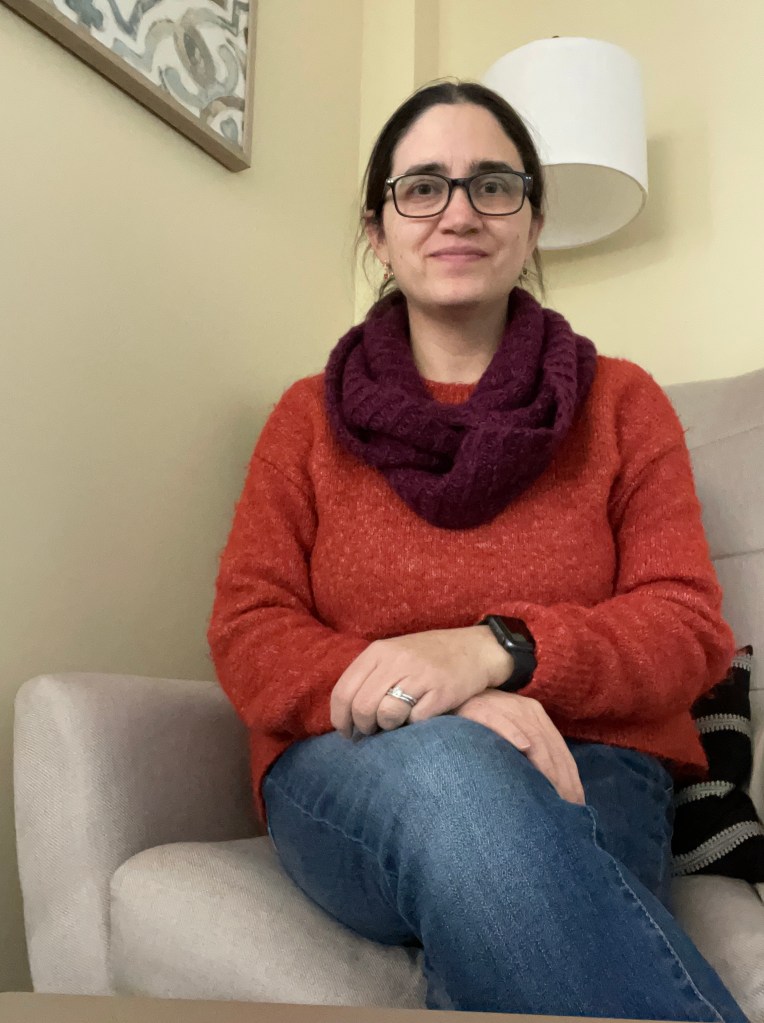 A friendly photo of Sarah in an orange sweater, purple scarf, and jeans, sitting in an office chair  
