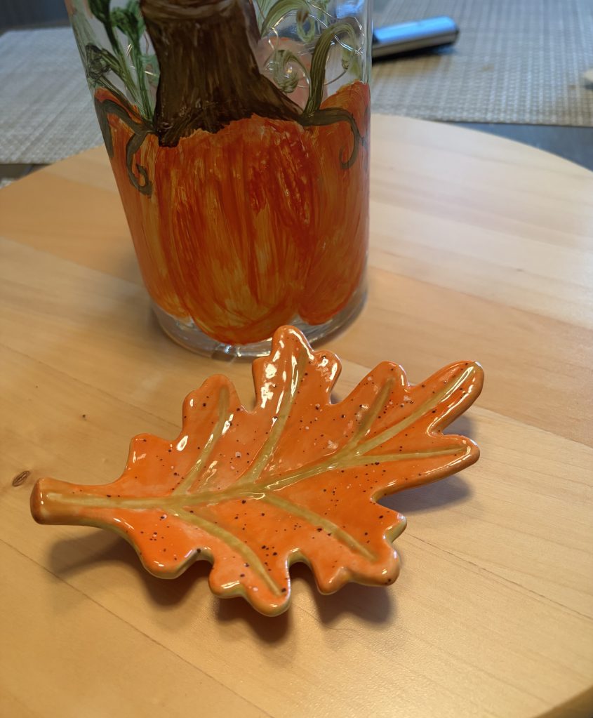 An orange ceramic leaf dish placed on a wooden table, next to a glass decorated with a painted pumpkin and vine design.